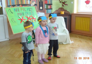 Troje dzieci z opaskami na głowie stoi w rzędzie i recytuje wiersz. Za nimi plakat "Nie pal przy mnie proszę".